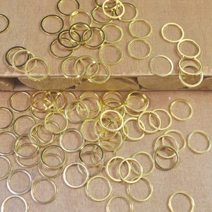 May include: A large pile of gold jump rings, a type of jewelry finding, are scattered on a wooden surface. The rings are various sizes and are perfect for crafting jewelry.