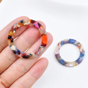 Puede incluir: Dos pendientes redondos de acrílico coloridos con un patrón moteado. Los pendientes son una mezcla de naranja, rojo, azul, blanco y marrón.