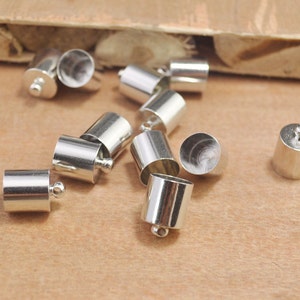 May include: A group of silver metal end caps for jewellery making. The end caps are cylindrical with a small ball on the end.