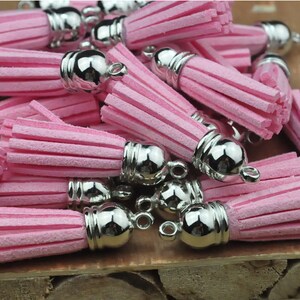 May include: A group of pink suede tassels with silver metal caps and loops. The tassels are arranged in a pile on a wooden surface.