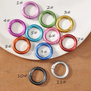 Può includere: Una collezione di undici anelli a scatto in metallo colorato, in varie tonalità, dal rosa all'argento. Ogni anello è numerato da 1 a 11. Gli anelli sono disposti su un piatto bianco e una superficie marrone. Adatti per progetti creativi.