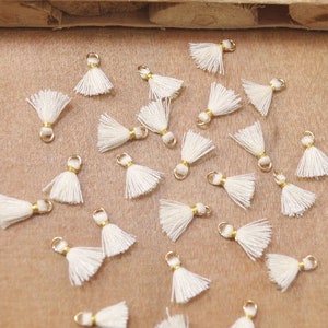 Op de afbeelding: Een verzameling witte kwastcharms met gouden springringen. De kwasten zijn gerangschikt op een lichtbruin oppervlak.