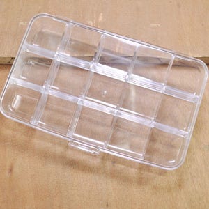May include: A clear plastic storage box with 12 compartments. The box is empty and has a slightly curved shape.