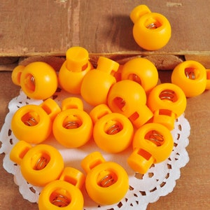 May include: A collection of 15 bright yellow plastic toggle clasps with a spring mechanism. The clasps are arranged in a circle on a white doily.