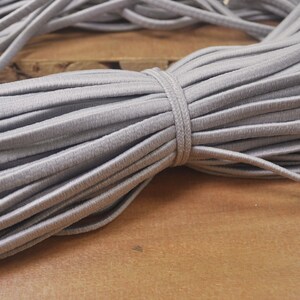 May include: A close-up of a spool of grey elastic cord. The cord is tightly wound and has a smooth, matte finish.