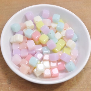 May include: A white bowl filled with pastel-colored square beads. The beads are in shades of pink, yellow, blue, and white. The beads have a hole through the center, suggesting they are for jewelry making or crafts.