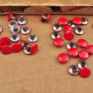 Puede incluir: Una colección de remaches metálicos rojos con tapas plateadas. Los remaches están dispersos sobre una superficie de madera.