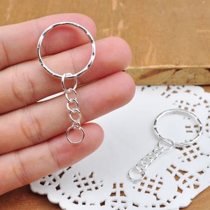 Puede incluir: Dos llaveros plateados con un anillo retorcido y una cadena plateada. Los llaveros están sobre un encaje blanco.
