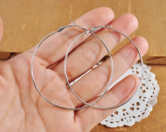 May include: A pair of silver hoop earrings. The earrings are large and circular, and they are shown on a person's hand.