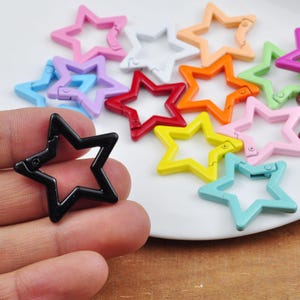 Puede incluir: Una colección de cierres en forma de estrella de colores, incluyendo negro, rojo, amarillo y azul. Los cierres están hechos de un material brillante y están dispuestos sobre una superficie blanca. Los cierres están diseñados para abrirse y cerrarse.