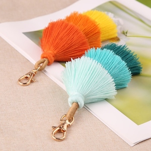 May include: Two colorful tassel keychains with gold metal clasps. The keychain on the left has orange, yellow, and red tassels. The keychain on the right has light blue, turquoise, and dark blue tassels.