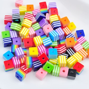 Puede incluir: Una colección de cuentas cuadradas de colores, con rayas. Las cuentas son de plástico y tienen una variedad de colores, incluyendo rojo, azul, verde, amarillo, naranja, morado y blanco.
