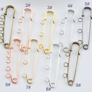 May include: Nine metal safety pins in various colors, including gold, silver, and bronze. Each pin has a loop or multiple loops for attaching beads or charms. The pins are numbered 1 through 9.