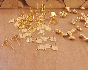 May include: A collection of gold-colored earring posts and backs. The posts have a flat, round base with a small loop and a straight post for attaching earrings. The earring backs are cylindrical with a rounded top.