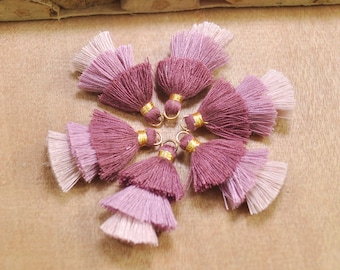 May include: Six pink and burgundy ombre tassel charms with gold accents. The tassels are arranged in a circle on a wooden surface.
