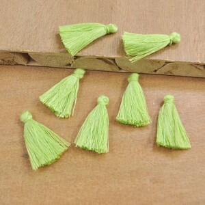 May include: Eight green tassel trims, each with a loop at the top, are scattered on a wooden surface. The tassels are made of soft, fluffy yarn and are approximately 1 inch in length.