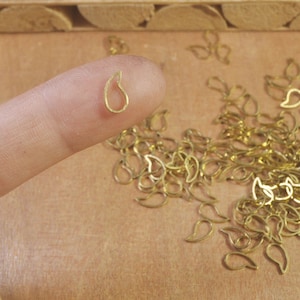 May include: A close-up of a small, gold-colored teardrop-shaped pendant. The pendant is held between a finger and thumb. The background is a pile of similar pendants.