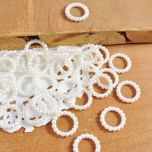 Puede incluir: Un montón grande de anillos de perlas blancas sobre un encaje blanco. Los anillos están hechos de pequeñas perlas blancas y están dispuestos en un patrón aleatorio.