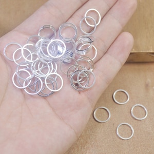 May include: A collection of small, silver-colored open rings. The rings are scattered across a light-colored surface, with some overlapping. The rings appear to be made of metal and are uniform in size and shape. They are likely jewelry components or craft supplies.