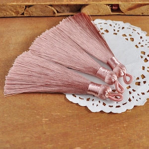 May include: Three pink silk tassels with a fringe of threads. The tassels are tied together and are on a white doily.