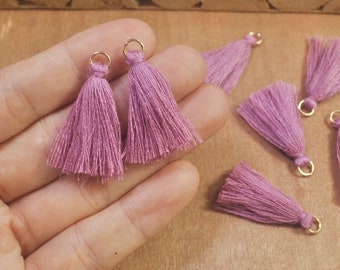 May include: A close-up of a hand holding two pink tassel charms with gold jump rings. The tassels are made of soft, fluffy yarn and are perfect for adding a touch of whimsy to jewelry or crafts.