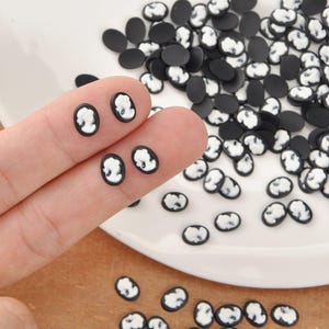 May include: Small, oval-shaped black and white cameo beads. The beads feature a white profile against a black background. Some beads are on a white plate, and some are held by a finger.
