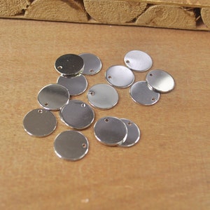 Op de afbeelding: Een groep van 15 zilverkleurige metalen cirkels met een klein gaatje in het midden. De cirkels liggen verspreid op een houten oppervlak.