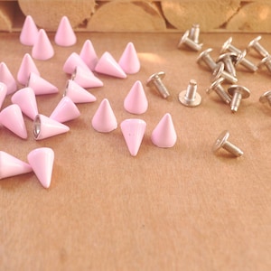 May include: A collection of pink metal pyramid studs and silver metal screw-back studs. The studs are arranged on a wooden surface.