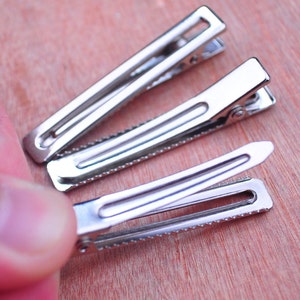 Op de afbeelding: Drie zilveren metalen haarclips worden getoond op een houten oppervlak. De clips zijn open en onthullen hun binnenwerk. De clips hebben een gekartelde rand en een scharnierend ontwerp voor een veilige haarstyling. De achtergrond is een warm, lichtbruin.
