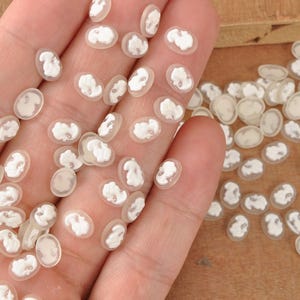 May include: A collection of small, oval-shaped cameo cabochons. Each cabochon features a white relief portrait set against a translucent background. The cabochons are scattered across a surface, showcasing their delicate details and vintage aesthetic.