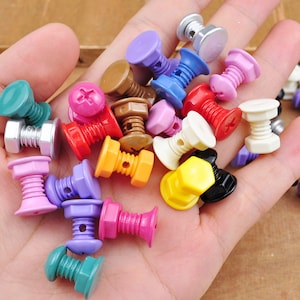 May include: Assortment of colorful plastic screw-shaped buttons in various colors, including pink, blue, green, yellow, white, and black. The buttons are arranged on a hand.