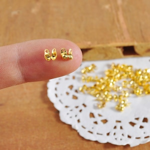 May include: Gold-colored metal clasps for jewelry making. Two clasps are shown on a finger, and a pile of clasps is shown on a white doily.
