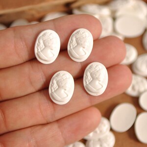May include: Four small, white oval cameos with a classical woman's profile in relief are held in a hand. The cameos are made of a smooth, matte material. Other cameos are visible in the background.