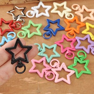 May include: A selection of star-shaped swivel clasps in a range of colours, such as red, green, blue, orange, yellow, pink, and purple. The clasps are made of a shiny material and have a metal ring. A black star clasp is held in a hand.