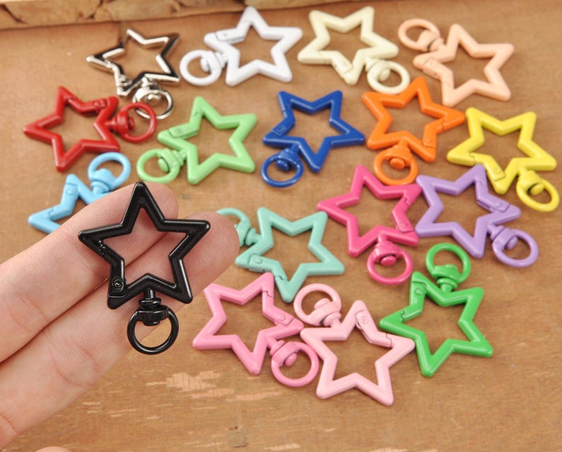 May include: A selection of star-shaped swivel clasps in a range of colours, such as red, green, blue, orange, yellow, pink, and purple. The clasps are made of a shiny material and have a metal ring. A black star clasp is held in a hand.