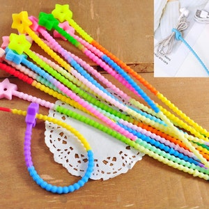 Puede incluir: Un conjunto de bridas para cables coloridas en varios tonos, incluyendo amarillo, verde, rosa y azul. Algunas bridas tienen extremos en forma de estrella, mientras que otras se utilizan para organizar cables. Las bridas están hechas de un material flexible y texturizado.