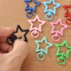 May include: A selection of star-shaped swivel clasps in colours such as black, blue, pink, and green. One black clasp is held by a hand. These clasps are likely used for jewellery making or crafting.