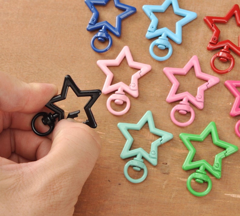 May include: A selection of star-shaped swivel clasps in colours such as black, blue, pink, and green. One black clasp is held by a hand. These clasps are likely used for jewellery making or crafting.