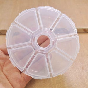 Może przedstawiać: Okrągłe pudełko na tabletki z przezroczystego plastiku z 10 przegródkami. Przegródki mają kształt trójkątów i są rozmieszczone w okręgu wokół centralnego otworu.