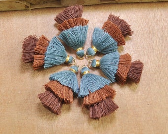 May include: A set of eight tassel trims in blue and brown. The tassels are made of two layers of fabric, with the top layer being blue and the bottom layer being brown. The tassels are attached to gold metal rings.