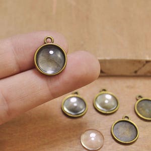 Może przedstawiać: Sześć okrągłych szklanych kaboszonów z antycznymi mosiężnymi oprawami. Kaboszony są przezroczyste i mają lekko wypukły kształt. Oprawy mają pętelkę na górze do przymocowania do łańcuszka lub sznurka.