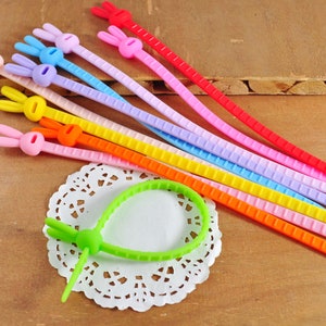 Może przedstawiać: Zestaw kolorowych opasek kablowych w kształcie królika w kolorach: czerwonym, różowym, fioletowym, jasnoniebieskim, niebieskim, żółtym, pomarańczowym i zielonym. Opaski są wykonane z elastycznego silikonu i mają fakturowaną powierzchnię.