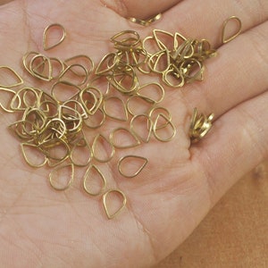 May include: A collection of small, gold-colored teardrop-shaped metal charms. The charms are open-backed and appear to be made of wire. They are scattered across a light brown surface, possibly a hand.