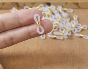 Puede incluir: Un conector de cadena de gafas de plástico blanco con un anillo de metal dorado. El conector está en la mano de una persona. Hay muchos más conectores en el fondo.