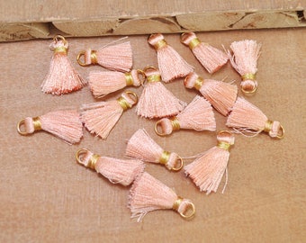 May include: A collection of 15 peach-colored tassels with gold wire accents. The tassels are arranged on a wooden surface.