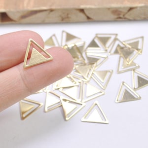 Peut inclure: Un tas de breloques triangulaires en métal doré avec un double contour. Les breloques sont éparpillées sur une surface blanche.