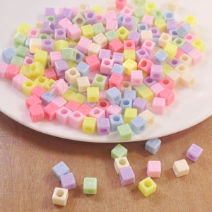 Könnte beinhalten: Eine Sammlung von pastellfarbenen Plastikbuchstabenperlen in verschiedenen Farben, darunter Rosa, Blau, Gelb und Grün. Die Perlen sind auf einem weißen Teller verstreut und einige liegen auf einer Holzoberfläche.