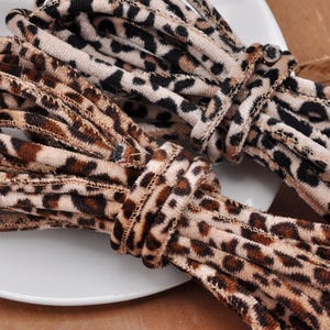 May include: Two bundles of cord featuring a soft, velvet-like texture with a brown, black, and beige leopard print pattern. The cords are coiled and display a consistent animal-inspired design across the entire length of the material.