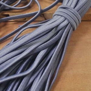May include: A bundle of grey elastic cord, also known as shoelace elastic, is shown on a wooden surface. The elastic is tightly wound together, creating a thick, rope-like appearance.