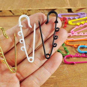Könnte beinhalten: Vier Sicherheitsnadeln in verschiedenen Farben: Gold, Weiß, Schwarz und Silber. Die Nadeln sind auf einer Hand angeordnet, wobei die goldene Nadel links und die silberne Nadel rechts ist.